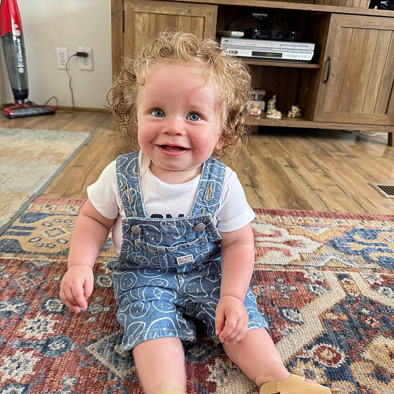 Timothy G is registered to the contest to win money with this photo: blue_eyes, casual_clothing, child, curly_hair, cute, denim_overalls, flooring, footwear, happy, home_decor, indoor, living_room, person, rug, sitting, smiling, television, toddler, wooden_cabinet, young_child