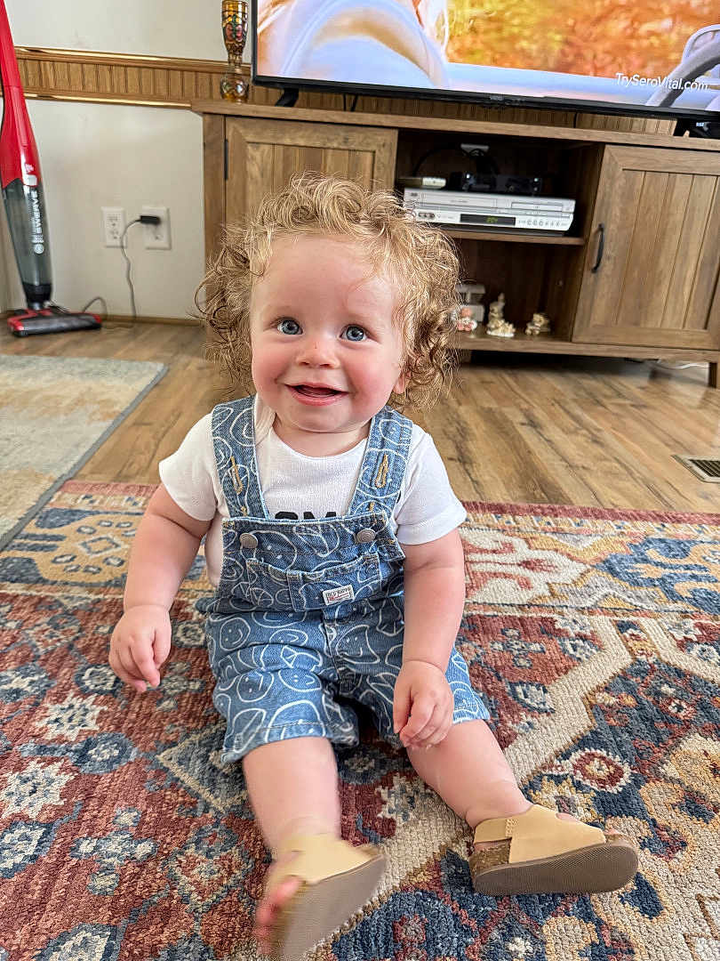 Timothy G is registered to the contest to win money with this photo: blue_eyes, casual_clothing, child, curly_hair, cute, denim_overalls, flooring, footwear, happy, home_decor, indoor, living_room, person, rug, sitting, smiling, television, toddler, wooden_cabinet, young_child