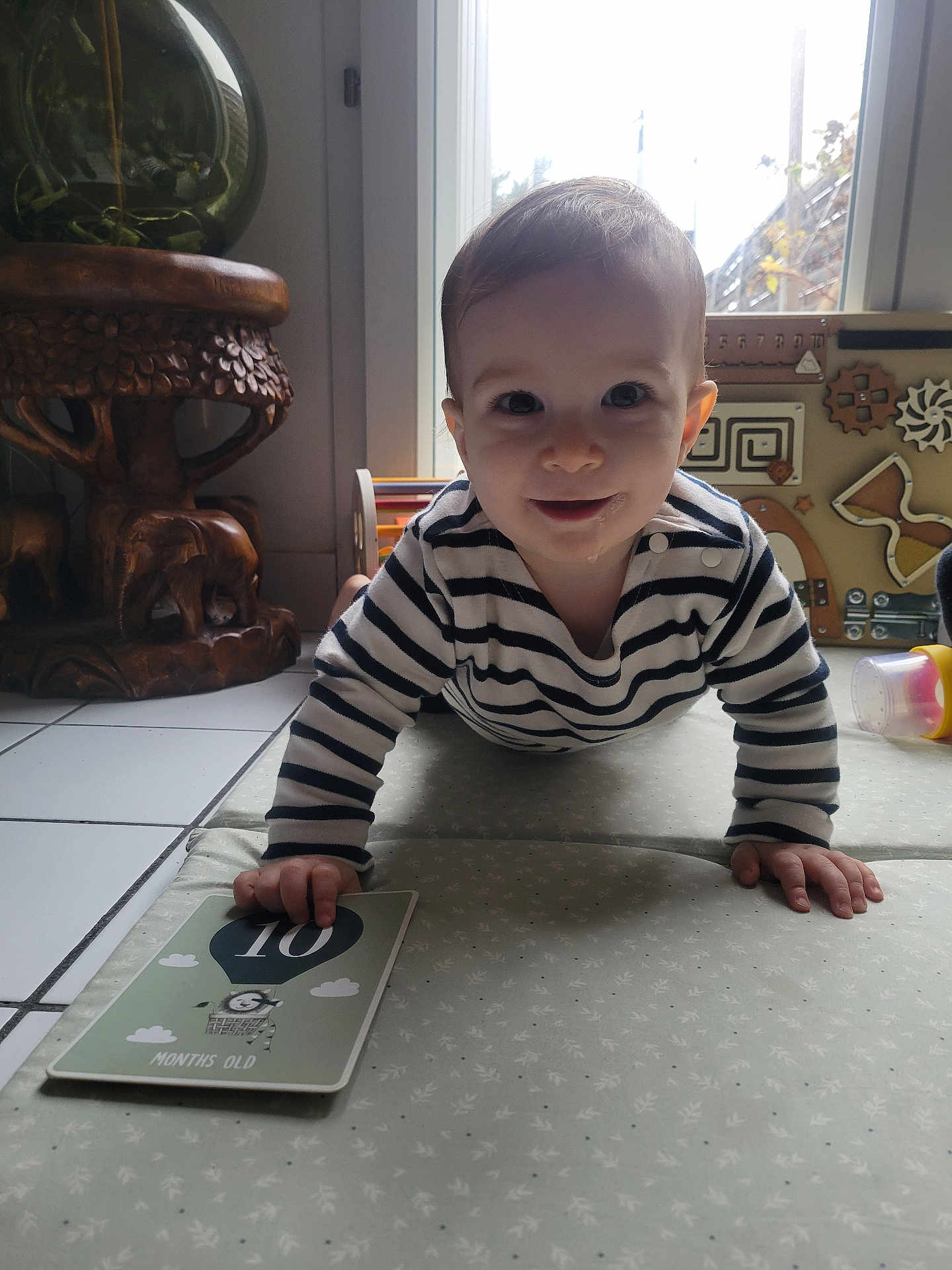 Adam a rejoint le concours — aidez-le/la à gagner de superbes lots ! baby, child, crawling, milestone_card, striped_clothing, drooling, indoor, soft_mat, toy, wooden_table, window, natural_light, floor_tiles, smiling, person, head, hand, face, home, cute