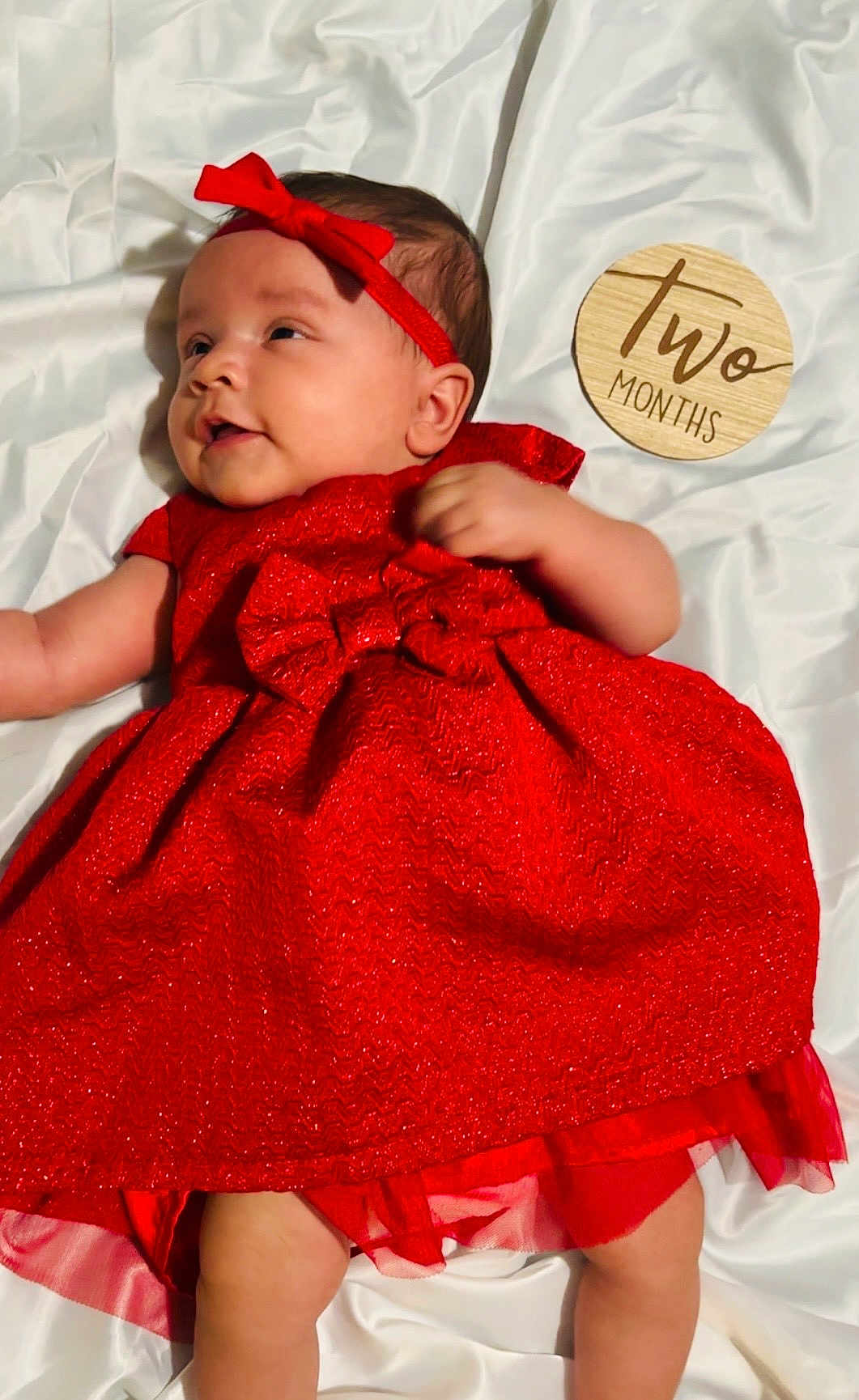 Angel is registered to the contest to win money with this photo: baby, infant, red_dress, headband, lying_down, white_sheet, milestone_sign, two_months, cute, smiling, child, portrait, soft_lighting, indoors, clothing, fabric, skin, baby_girl, celebration, adorable