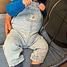 baby, pacifier, blue_overalls, socks, indoor, child, person, cushion, chair, hand, jeans, face, seated, cute, infant, holding, clothing, wooden_furniture, expression, closeup