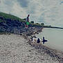 dog, person, lake, rocks, grass, cloudy_sky, water, shore, beach, outdoor, nature, footwear, green_shorts, black_and_white_dog, walking, landscape, recreation, summer, casual_clothing, daytime