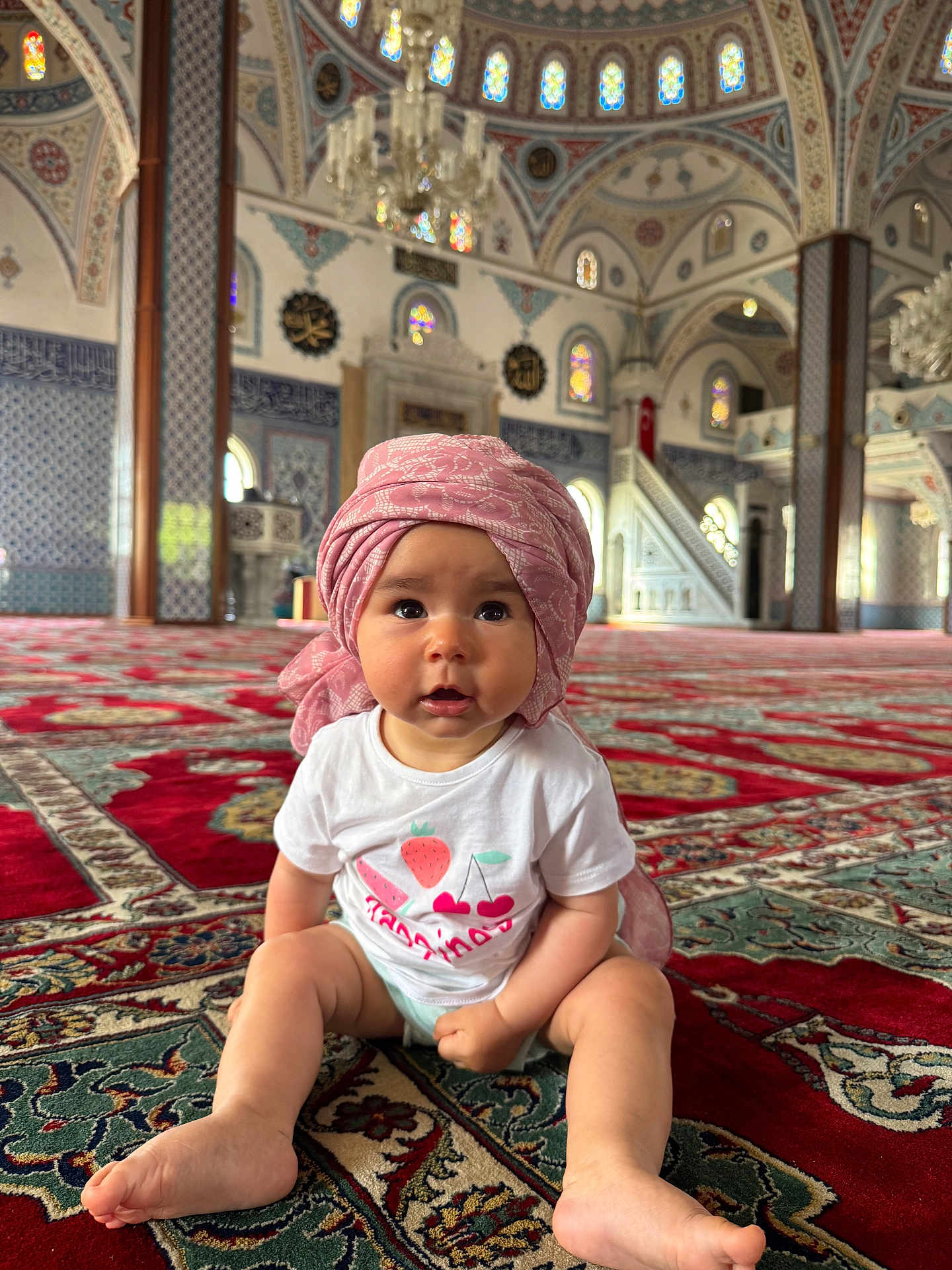 Aelis a rejoint le concours — aidez-le/la à gagner de superbes lots ! baby, child, indoor, mosque, carpet, turban, white_shirt, fruit_graphics, architecture, stained_glass, patterned_floor, colorful, sitting, curious, portrait, person, headwear, decorative, religious_building, soft_lighting