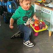 Eden a rejoint le concours — aidez-le/la à gagner de superbes lots ! child, toy_food, play_kitchen, toy_tractor, indoor, green_shirt, jeans, sneakers, floor, play, pretend_play, concentration, tongue_out, tray, furniture, cabinet, stove, knobs, toy, childhood
