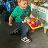 child, toy_food, play_kitchen, toy_tractor, indoor, green_shirt, jeans, sneakers, floor, play, pretend_play, concentration, tongue_out, tray, furniture, cabinet, stove, knobs, toy, childhood