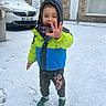 child, snow, winter_clothing, jacket, scarf, hood, sneakers, footprints, car, building, stone_wall, door, window, outdoor, playful, smiling, cold_weather, street, person, casual_wear