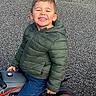 child, boy, smile, green_jacket, jeans, toy_bike, orange_wheels, outdoor, gravel, happy, portrait, casual_clothing, play, vehicle, childhood, fun, young, cute, person, joy