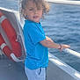 blue_shirt, boat, boy, casual_clothing, child, curly_hair, daylight, deck, life_saver, nautical, ocean, outdoor, portrait, railing, shorts, standing, summer, sunlight, vacation, water