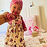 Janelis is registered to the contest to win money with this photo: baby, child, smiling, tongue_out, headband, pink, dress, floral, bed, bedding, plush_toy, hand_holding, person, indoors, cute, happy, infant, pillow, toy, sleepwear