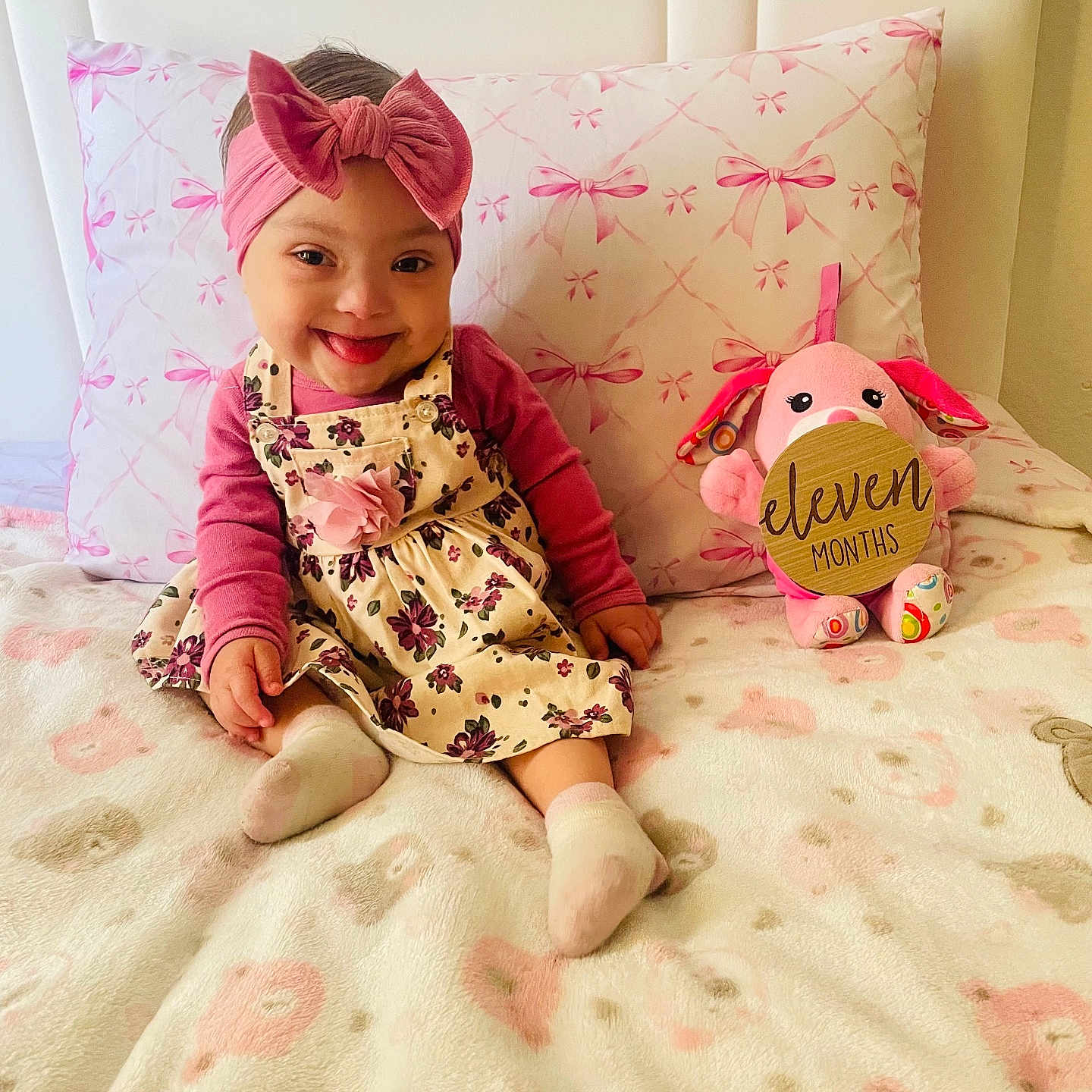 Janelis joined the competition — help win amazing prizes! baby, child, smile, headband, bow, dress, floral, pink, plush_toy, bed, blanket, pillow, indoor, cute, sitting, happy, person, infant, sign, soft_toy