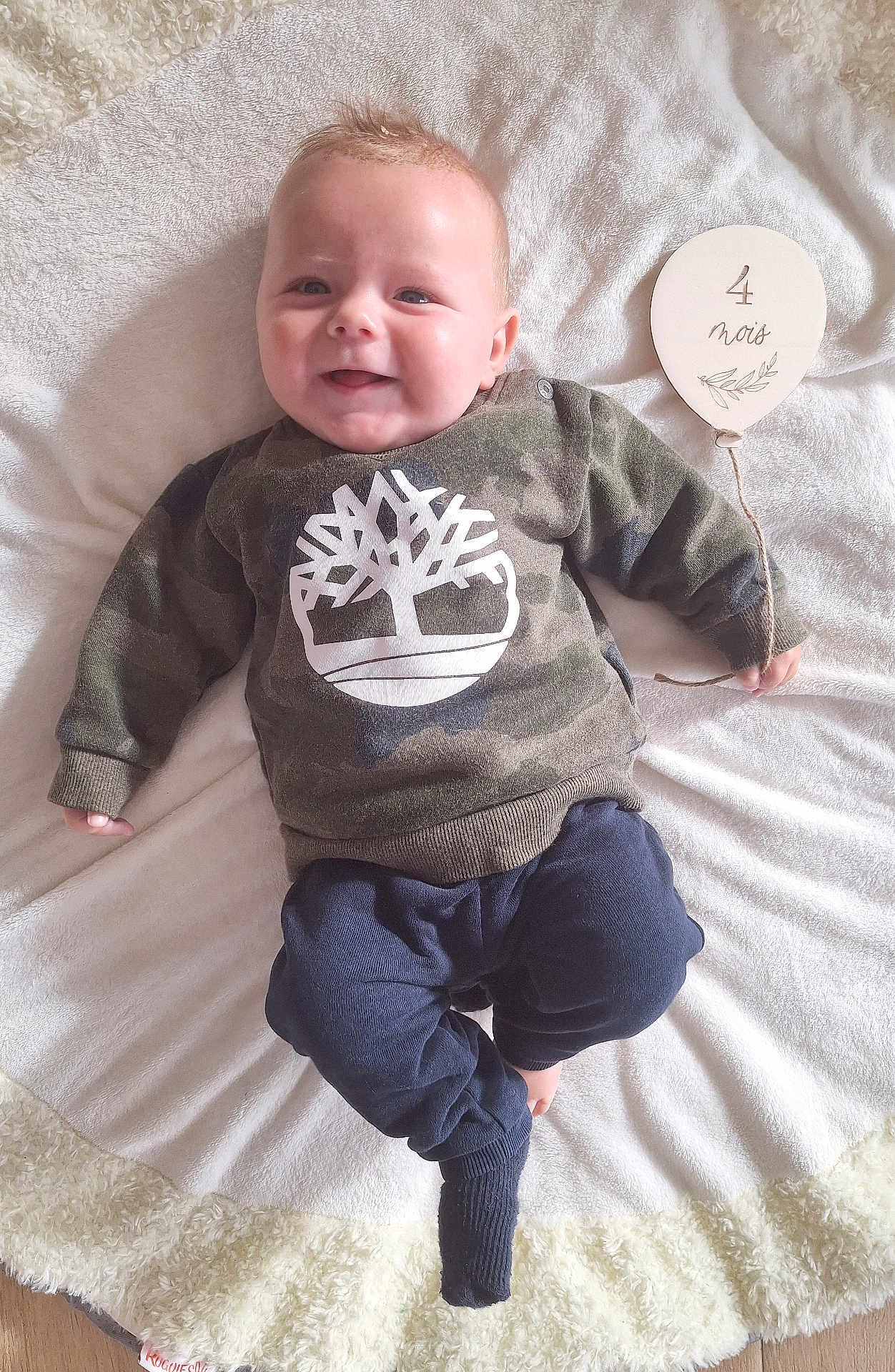 Menzio participe au concours pour gagner de l'argent avec cette photo : baby, infant, smiling, camo_sweater, navy_pants, blanket, wooden_sign, balloon_shape, four_months, lying_down, cute, happy, indoors, soft_texture, child, person, clothing, face, hand, foot