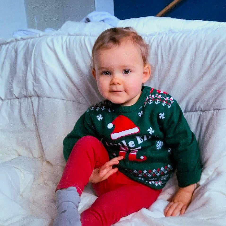 Lisa a rejoint le concours — aidez-le/la à gagner de superbes lots ! bed, blanket, child, christmas, cozy, cute, face, furniture, holiday, home, indoors, portrait, red_pants, relaxed, smile, socks, sweater, toddler, white_bedding, young_child