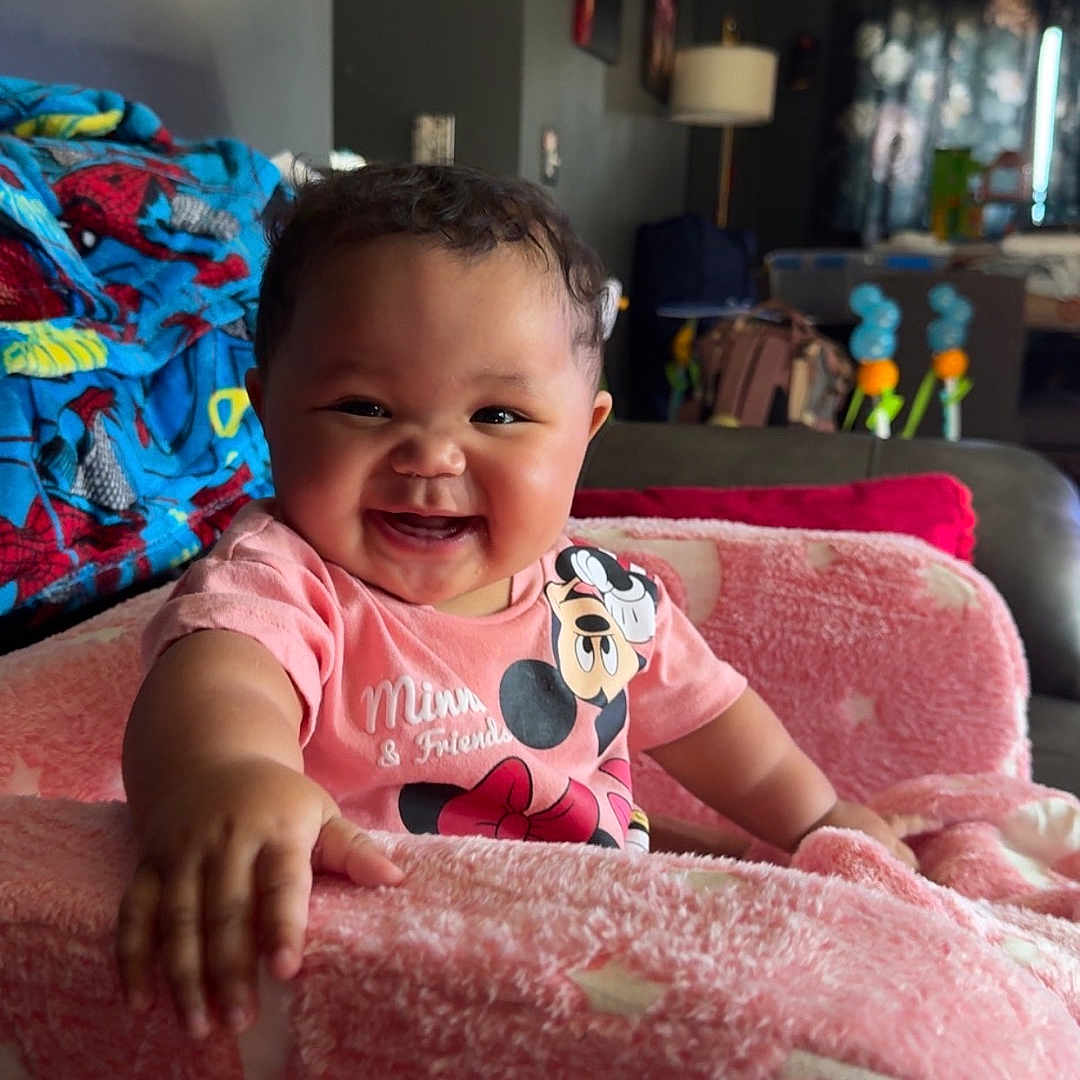 Araya joined the competition — help win amazing prizes! baby, smiling, pink_chair, blanket, indoor, child, happy, cozy, living_room, furniture, toddler, curly_hair, person, soft_texture, cute, seated, casual_clothing, home, colorful, comfort
