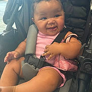 Araya is registered to the contest to win money with this photo: baby, child, stroller, pink_clothing, smile, happy, infant, seatbelt, purse, cute, indoors, chubby_cheeks, person, young_child, sitting, hand, foot, black_stroller, casual, relaxed