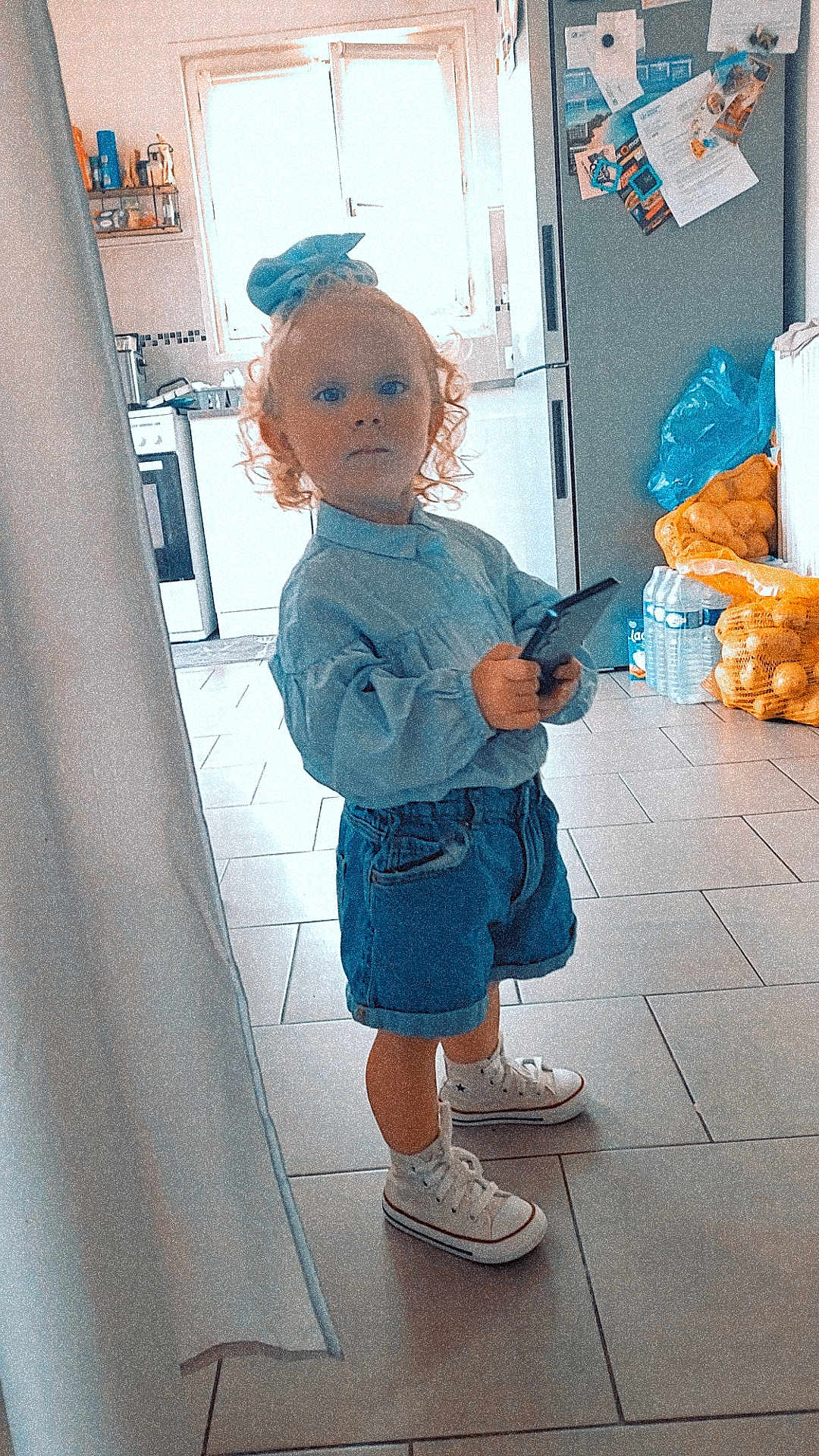 Milla a rejoint le concours — aidez-le/la à gagner de superbes lots ! toddler, child, curly_hair, denim_shorts, white_sneakers, remote_control, kitchen, fridge, notes, magnets, window, tile_floor, plastic_bags, potatoes, water_bottles, blue_bow, casual_clothing, standing, indoor, person