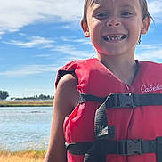 Phoenix is registered to the contest to win money with this photo: child, smile, life_jacket, red, outdoor, water, lake, sky, clouds, sunlight, summer, nature, happy, portrait, boy, teeth, grass, daylight, person, vacation