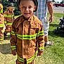 child, firefighter, costume, helmet, smiling, outdoor, grass, person, adult, boots, sunny, daylight, uniform, event, people, fun, park, family, celebration, safety