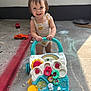 toddler, child, toy, push_toy, floor, chalk_drawing, outdoor, smile, happy, clothing, plastic, wheels, toy_truck, curb, concrete, sunlight, play, person, colorful, background
