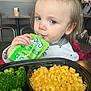 toddler, child, food_pouch, broccoli, macaroni_and_cheese, table, restaurant, indoor, person, face, blue_eyes, hair, clothing, seat, meal, plate, eating, curious, casual_dining, background