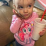 toddler, child, pink_shirt, my_little_pony, cup, straw, phone, indoor, carpet, couch, blanket, papers, curious_expression, standing, hair_buns, casual_clothing, person, drink, toy, home