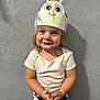 Josie is registered to the contest to win money with this photo: toddler, child, paper_crown, smile, happy, standing, short_sleeves, white_shirt, black_and_white_pants, indoor, wall, texture, hands, face, cute, playful, young_child, expression, portrait, casual_clothing