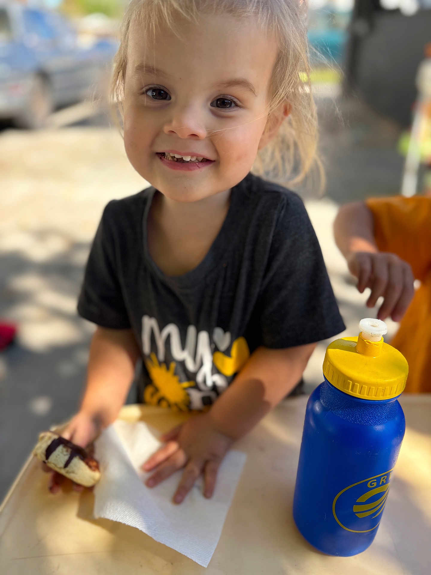 Josie is registered to the contest to win money with this photo: child, toddler, smiling, face, hand, napkin, food, cookie, water_bottle, table, outdoor, daylight, clothing, short_sleeve, yellow, blue, happy, person, closeup, blurred_background