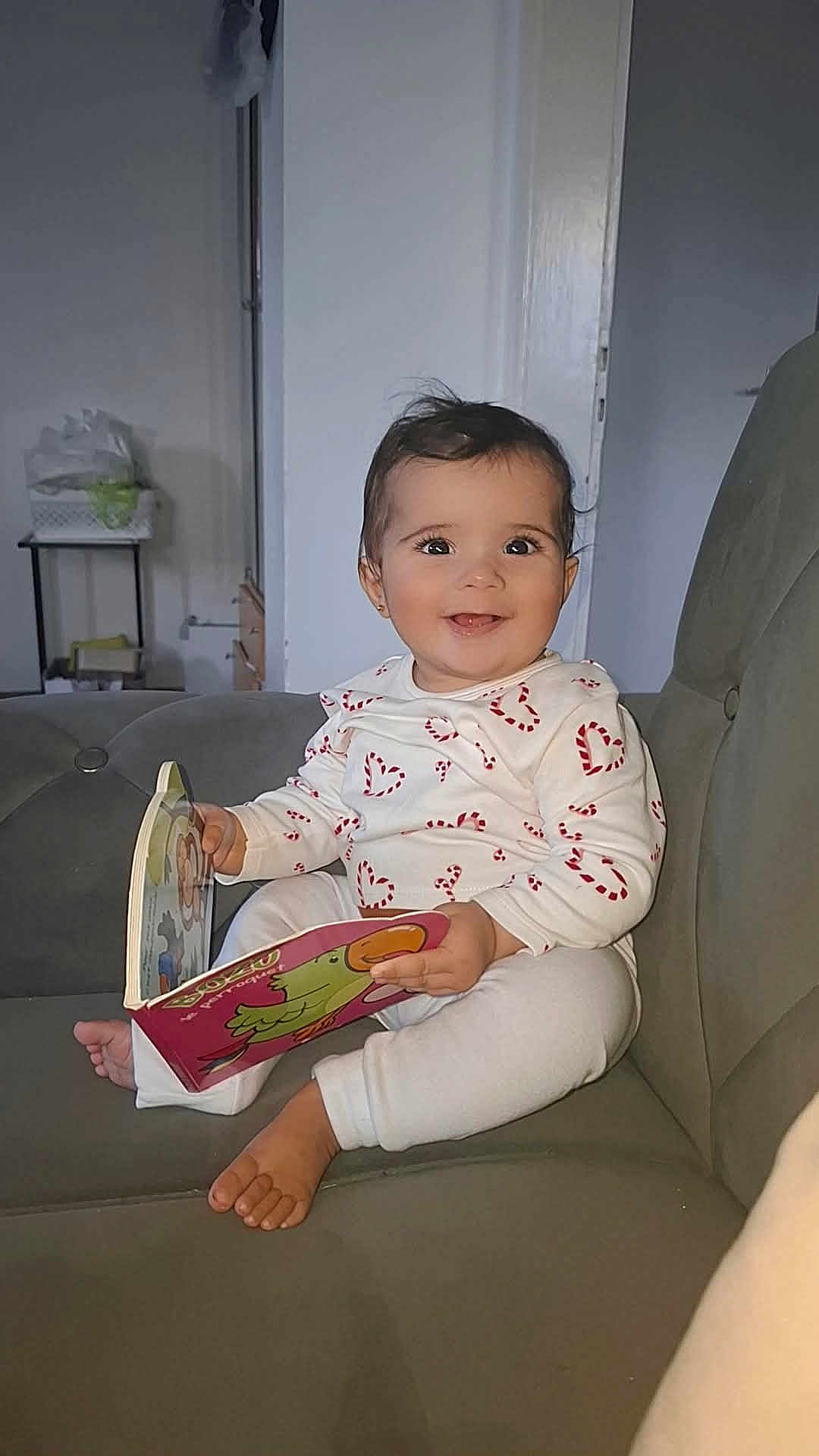 Nour a rejoint le concours — aidez-le/la à gagner de superbes lots ! baby, child, smiling, reading, book, couch, indoor, white_clothing, heart_pattern, barefoot, happy, person, furniture, cute, infant, seated, toy, living_room, portrait, playful