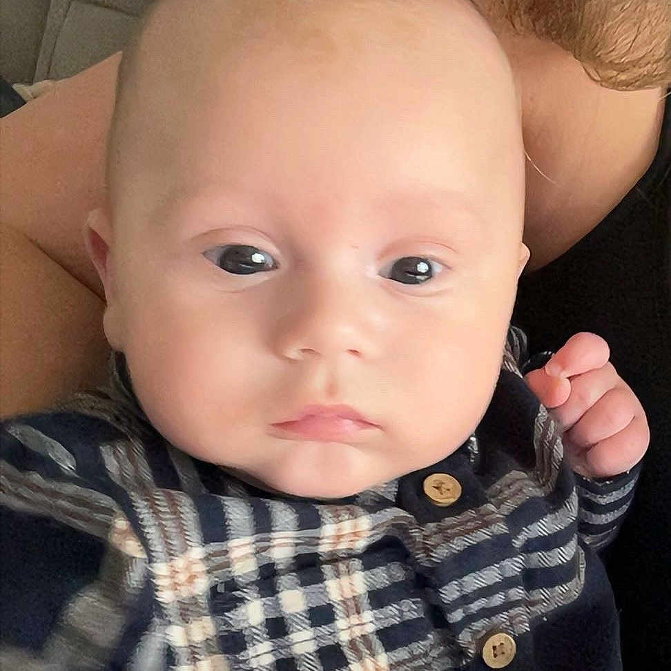 Soann participe au concours pour gagner de l'argent avec cette photo : adult_arm, baby, bald_head, calm_expression, cheeks, closeup, couch, eyes, face, hand, holding, indoor, infant, looking_at_camera, newborn, plaid_shirt, portrait, sitting, soft_skin, wooden_buttons