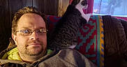 Licorice is registered to the contest to win money with this photo: beard, blanket, cat, couch, curtain, cushion, glasses, hoodie, indoor, living_room, man, patterned_blanket, pet, portrait, relaxed, seated, selfie, tabby_cat, window, wood_paneling