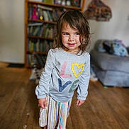 Ila is registered to the contest to win money with this photo: child, indoor, barefoot, sweater, colorful, love, striped_skirt, wooden_floor, bookshelf, couch, casual_clothing, portrait, smiling, tousled_hair, young_child, floor, person, home, cozy, cute