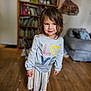child, indoor, barefoot, sweater, colorful, love, striped_skirt, wooden_floor, bookshelf, couch, casual_clothing, portrait, smiling, tousled_hair, young_child, floor, person, home, cozy, cute