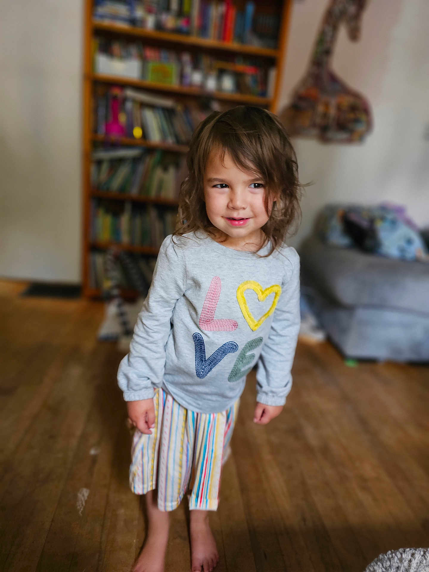 Ila is registered to the contest to win money with this photo: child, indoor, barefoot, sweater, colorful, love, striped_skirt, wooden_floor, bookshelf, couch, casual_clothing, portrait, smiling, tousled_hair, young_child, floor, person, home, cozy, cute