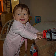 Lena participe au concours pour gagner de l'argent avec cette photo : baby, child, pink_onesie, smiling, toy_bus, wooden_table, indoor, happy, standing, playroom, furniture, toy, face, person, home, floor, expression, cute, infant, room