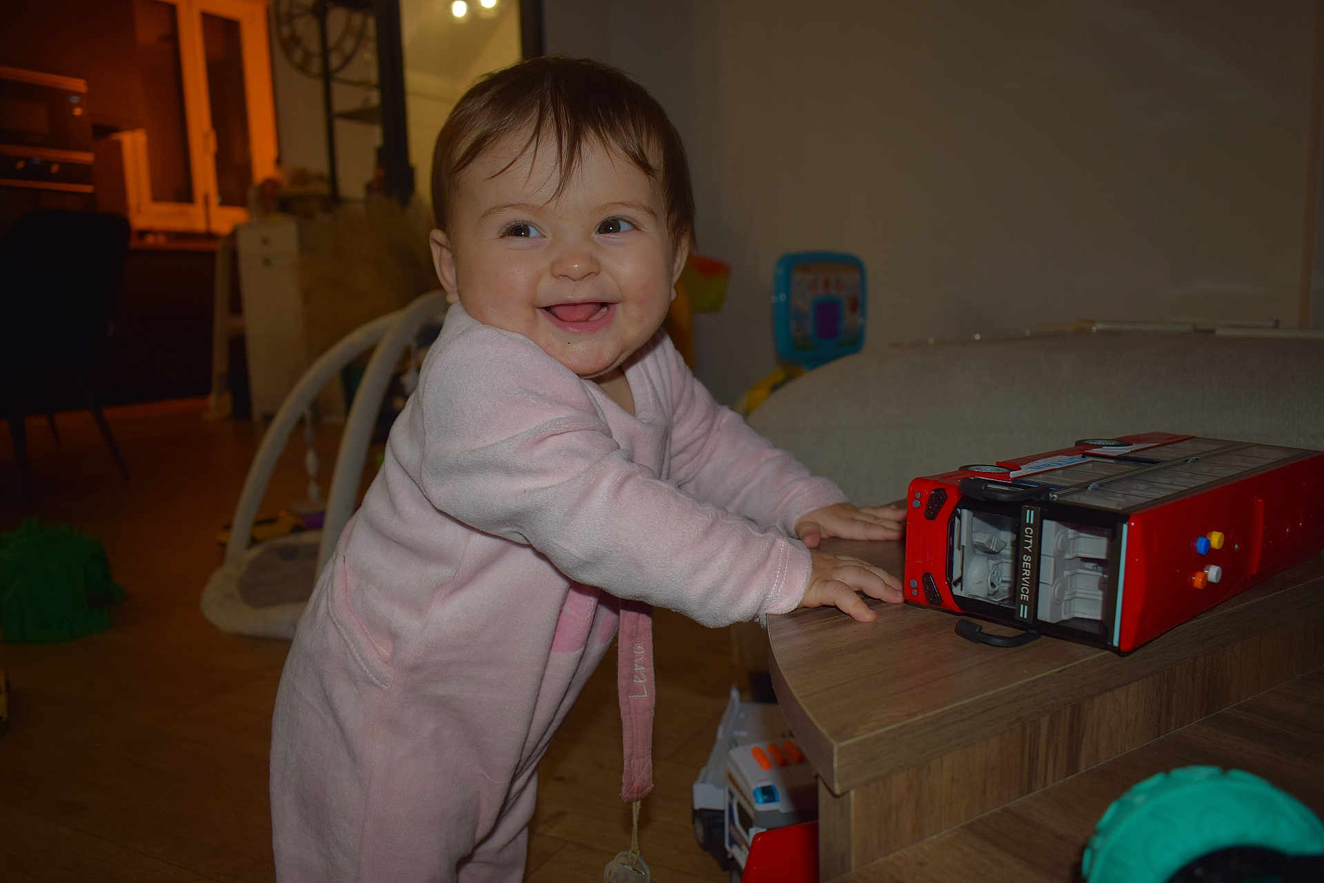 Lena participe au concours pour gagner de l'argent avec cette photo : baby, child, pink_onesie, smiling, toy_bus, wooden_table, indoor, happy, standing, playroom, furniture, toy, face, person, home, floor, expression, cute, infant, room