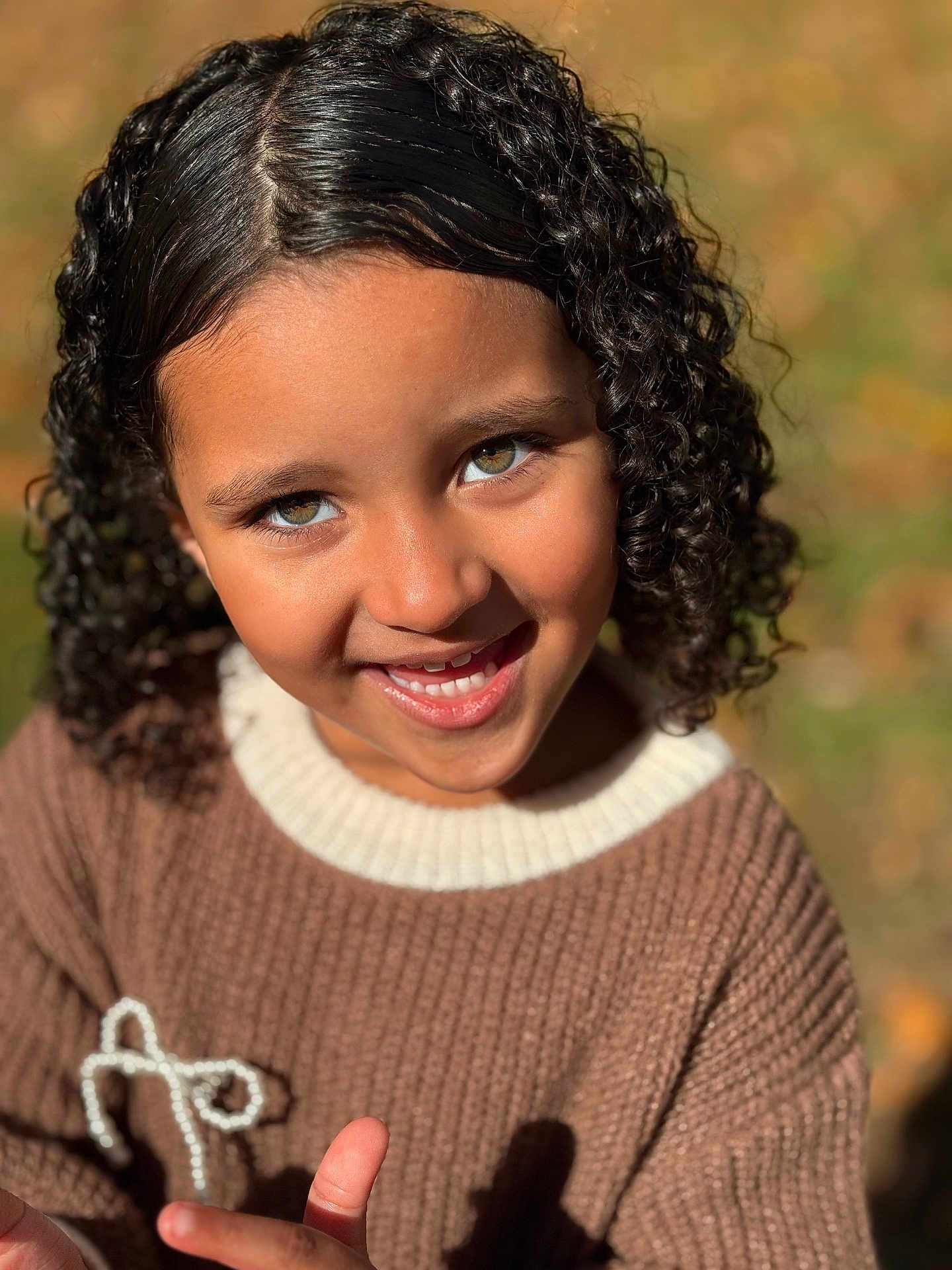 Laura is registered to the contest to win money with this photo: child, smiling, curly_hair, sweater, brown_clothing, outdoor, portrait, happy, face, person, childhood, sunlight, natural_light, close_up, youth, cute, warm_colors, fashion, fall, candid