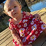toddler, child, dress, red_clothing, daisy_pattern, barefoot, wooden_dock, river, bridge, outdoor, sunlight, nature, water, reflection, trees, curly_hair, face, portrait, sitting, daytime