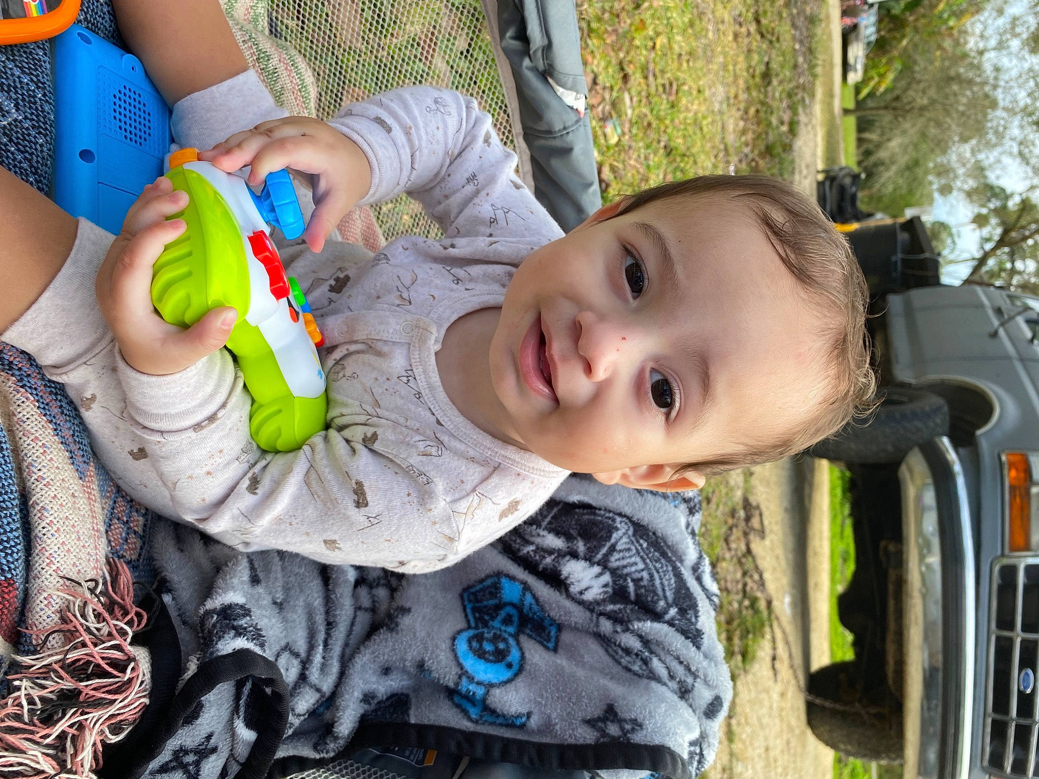 Elijah is registered to the contest to win money with this photo: automotive_exterior, baby, baby_products, child, fun, grass, happy, joy, leisure, motor_vehicle, pattern, person, plant, play, recreation, sitting, smile, toddler, toy, tree
