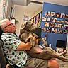man, dog, indoor, couch, colorful_shirt, glasses, family_photos, blue_wall, casual, relaxed, pet, home, decor, carpet, seated, holding, shorts, bracelets, living_room, portrait