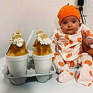 Tatum is registered to the contest to win money with this photo: baby, infant, pumpkin_outfit, orange_hat, shaved_ice, whipped_cream, cookie, cup, table, wall, power_outlet, hand_holding_baby, food, dessert, indoor, person, cute, snack, white_background, sitting