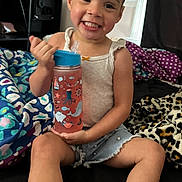 Miliani Rose joined the competition — help win amazing prizes! toddler, child, smiling, water_bottle, curly_hair, shorts, tank_top, bed, blanket, sneakers, indoor, happy, person, furniture, television, home, cute, playful, sitting, drink