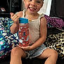 toddler, child, smiling, water_bottle, curly_hair, shorts, tank_top, bed, blanket, sneakers, indoor, happy, person, furniture, television, home, cute, playful, sitting, drink
