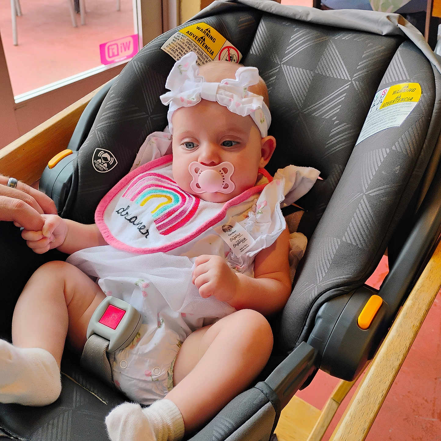 Ava is registered to the contest to win money with this photo: baby, infant, car_seat, pacifier, headband, bib, rainbow, hand_holding, indoor, child, cute, clothing, seatbelt, sock, person, portrait, wooden_furniture, fabric, safety, toddler