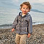 Sacha a rejoint le concours — aidez-le/la à gagner de superbes lots ! child, toddler, curly_hair, jacket, shorts, shoes, beach, pebbles, water, sea, outdoor, nature, person, standing, clothing, casual, young, cute, portrait, daylight