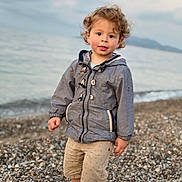 Sacha participe au concours pour gagner de l'argent avec cette photo : child, toddler, curly_hair, jacket, shorts, shoes, beach, pebbles, water, sea, outdoor, nature, person, standing, clothing, casual, young, cute, portrait, daylight