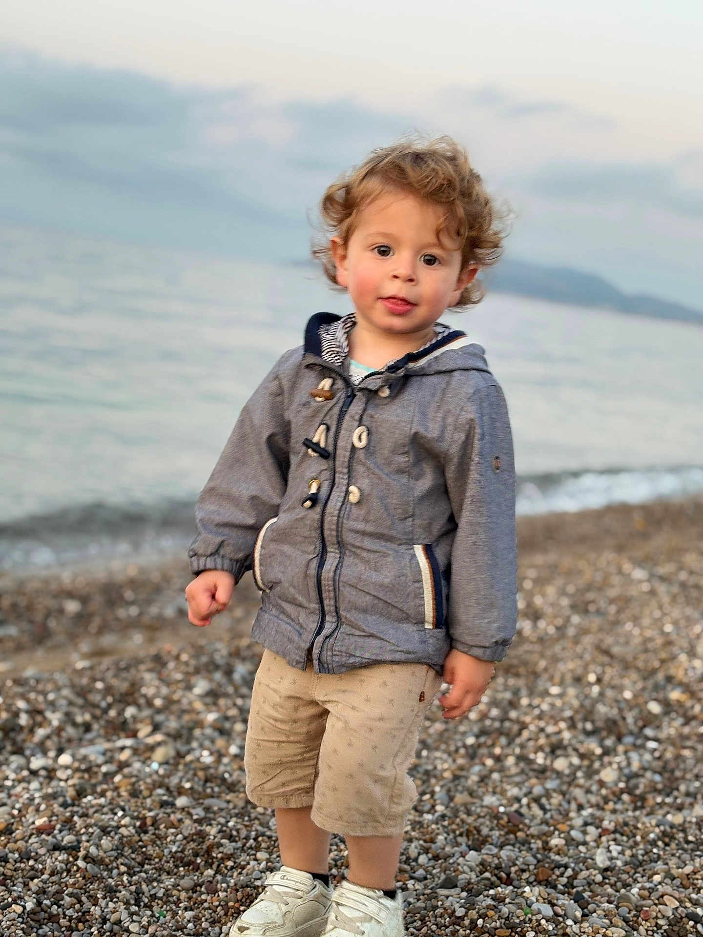 Sacha participe au concours pour gagner de l'argent avec cette photo : child, toddler, curly_hair, jacket, shorts, shoes, beach, pebbles, water, sea, outdoor, nature, person, standing, clothing, casual, young, cute, portrait, daylight