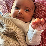 Zeyna Rose joined the competition — help win amazing prizes! baby, infant, blanket, knitted, cozy, smiling, face, hand, gesture, clothing, cushion, red, pattern, portrait, young_child, cute, indoors, soft_light, closeup, person