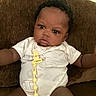 Amari is registered to the contest to win money with this photo: baby, child, infant, onesie, pacifier_clip, brown_couch, furniture, indoor, person, cute, curly_hair, expression, seated, portrait, young_child, casual, relaxed, skin, face, looking