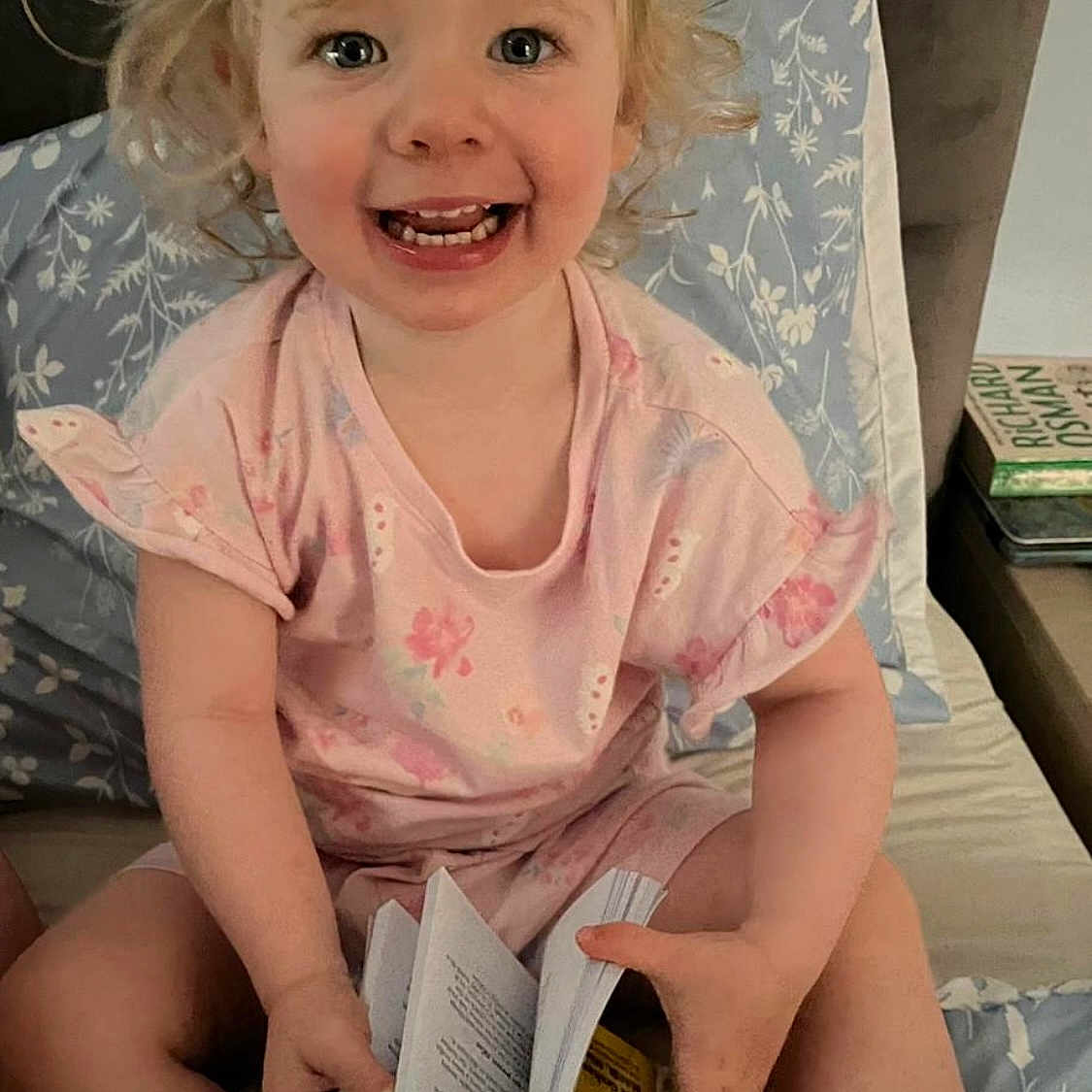 Sophia is registered to the contest to win money with this photo: bed, bedding, blonde_hair, book, casual_clothing, child, cozy, curly_hair, furniture, happy, home, indoor, person, pillow, pink_shirt, portrait, reading, smiling, table, toddler