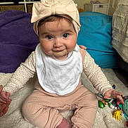 Frankie is registered to the contest to win money with this photo: baby, bib, blanket, bow, child, clothing, cushion, cute, expression, face, floor, hand, headband, indoor, infant, person, sitting, smile, sock, toy