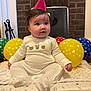 baby, party_hat, balloons, indoor, carpet, blanket, cushion, cute, child, white_clothing, celebration, birthday, infant, plush, face, sitting, floor, headwear, smiling, decor